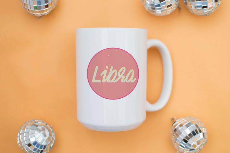 May include: White ceramic mug with a pink circle containing the word "Libra" in a cursive font. The mug is on an orange surface, with disco ball ornaments in the background.