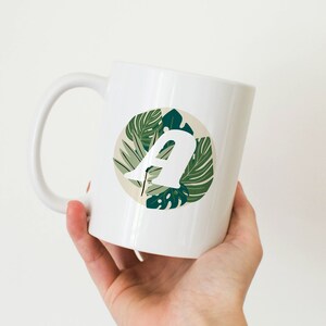May include: White ceramic mug with a circular design featuring the letter "A" in white, set against green tropical leaves. The mug has a curved handle and is held against a white backdrop.