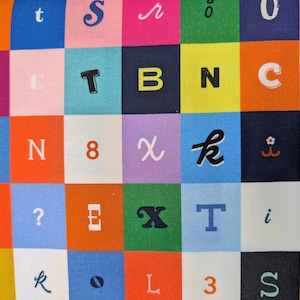 May include: A colorful fabric with a grid pattern featuring various letters, numbers, and symbols in a variety of fonts and colors. The squares are in shades of pink, blue, orange, green, and purple. The letters include T, B, N, C, X, and S.