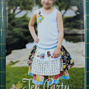 May include: A young girl wearing a white tank top with a floral embellishment and a colorful apron skirt. The skirt has multiple patterned layers. The text "Tea Party apron skirt and top" is visible. The image is from Pink Fig patterns.