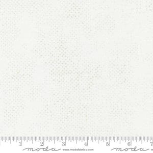 May include: A white fabric featuring a subtle, light gray speckled pattern across the surface. A ruler is visible at the bottom edge, showing measurements in inches, with the text 'moda' and 'www.modafabrics.com' printed below.