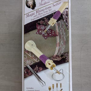 May include: A package for a Magic Rhinestone Setter and Mini Iron Set by Jenny Haskins. The set includes a support stand, tweezers, and various tips. The tool has a cream-colored handle with a purple grip. The box features a photo of a woman.