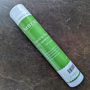 May include: A cylindrical roll of Kimberbell Fusible Backing for sewing and embroidery, featuring a bright green label with white text. The label specifies dimensions of 12 inches by 5 yards and includes a barcode and product information.