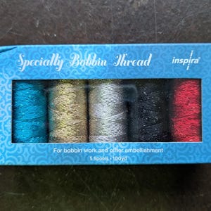 May include: A box of Inspira specialty bobbin thread in various colors. The box contains five spools of thread, including teal, gold, silver, black, and red. The box is blue with the words "Specialty Bobbin Thread" printed on it.