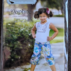 May include: A sewing pattern package for "Daphne" classic knickers. The package features a young girl wearing a white tank top and blue floral print knickers. The text on the package reads "Violette Field Threads" and "Sizes 2T-10 Years."