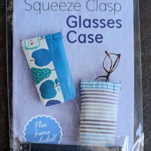 May include: A glasses case kit with the text "Squeeze Clasp Glasses Case". The kit includes two fabric cases, one with a blue apple pattern and the other with blue and brown stripes. A flex frame and hardware are included.