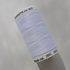 May include: A white spool of thread, labeled "BOBBINETTE Art. 0277" and "194 yds col. 2000 KSCK9." The thread is tightly wound, showing the texture of the fibers. Ideal for sewing, quilting, and other fabric crafts.