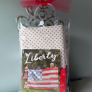 May include: A gift bag containing a quilt kit. The bag is clear with a red bow and handle. Inside, a fabric with red and black polka dots, a quilt pattern book titled "Liberty", and a red fabric with white stars are visible. The quilt pattern book shows a quilt designed as an American flag.