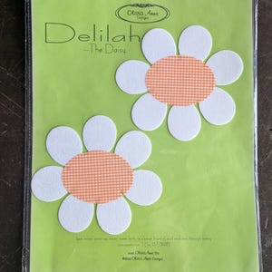 May include: Two daisy-shaped craft embellishments. Each daisy has white petals and an orange gingham center. The packaging is green and features the text "Delilah - The Daisy" and the Olivia Anne Designs logo.