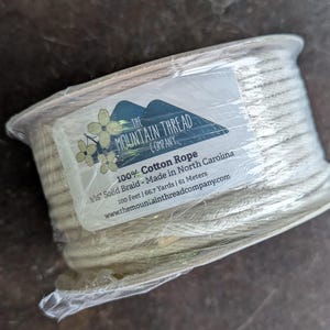 May include: A spool of off-white cotton rope, wrapped in clear plastic. The label reads "The Mountain Thread Company" and "100% Cotton Rope." The rope is 3/16" solid braid, 200 feet long, or 66.7 yards, and 61 meters.