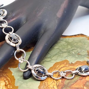 Op de afbeelding: Een zilveren armband met zwarte ovale stenen en kleine bloem accenten. De armband ligt op een hand en een getextureerd oppervlak.