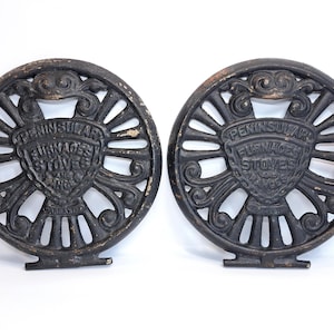 May include: Two circular black cast iron stove grates featuring an ornate openwork design with radiating spokes. Each piece displays a central shield embossed with the text PENINSULAR FURNACES STOVES AND RANGES, showing signs of aged wear.