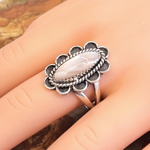 May include: A silver ring with an oval, light pink stone, surrounded by a decorative silver border. The ring has a floral-like design around the stone and a split band.