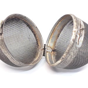 May include: Open tea infuser with a mesh basket and a hinged, metal frame. The infuser is open, revealing the fine mesh interior. The metal frame has a worn, antique appearance.