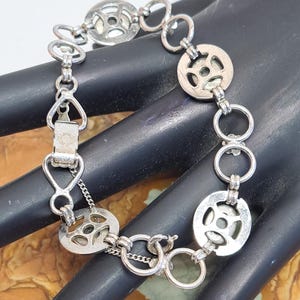 Op de afbeelding: Een zilveren armband met een kettinglinkontwerp en ronde bedels. De bedels hebben een uitgesneden patroon en zijn met kleine ringen aan de ketting bevestigd.