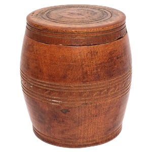 May include: A small, wooden barrel-shaped container with a lid. The container is made of a warm, reddish-brown wood and features horizontal grooves and decorative carvings. The lid has a circular design carved into its surface.