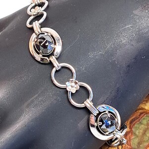 Op de afbeelding: Een zilveren armband met zwarte edelsteenaccenten. De armband heeft een kettinglinkontwerp met ronde, open cirkels en ovale edelstenen die in zilver zijn gezet.