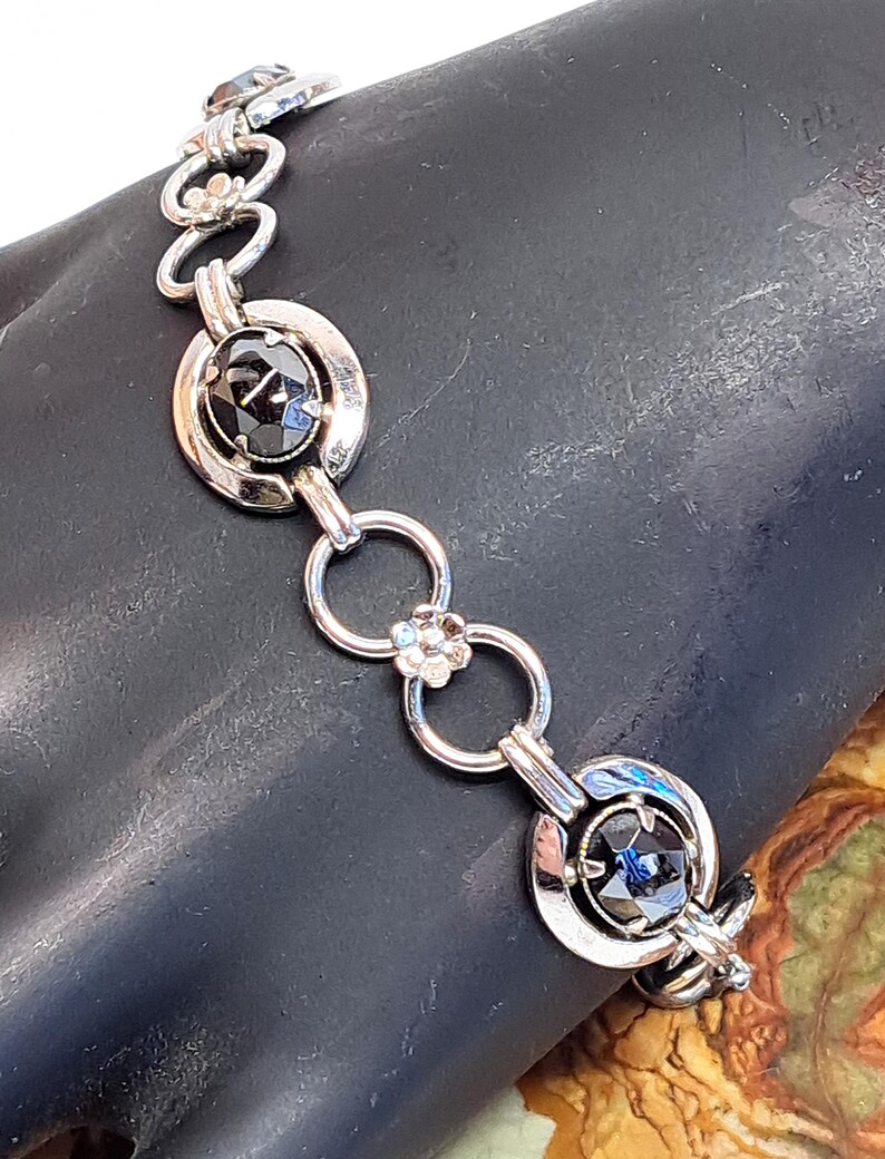 Op de afbeelding: Een zilveren armband met zwarte edelsteenaccenten. De armband heeft een kettinglinkontwerp met ronde, open cirkels en ovale edelstenen die in zilver zijn gezet.