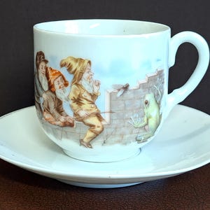 May include: White porcelain teacup and saucer set. The cup features a whimsical illustration of gnomes, a frog, and a dragonfly. The saucer has a simple, elegant design. A charming addition to any teacup collection.