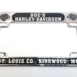 May include: Chrome license plate frame with the text "DOC'S HARLEY-DAVIDSON" in white on a black background. The frame also includes the text "ST. LOUIS CO., KIRKWOOD, MO". Harley-Davidson logos are on the top corners.