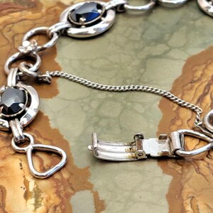 Op de afbeelding: Een zilveren armband met zwarte ovale stenen en een zilveren sluiting. De armband ligt op een bruine en witte achtergrond.