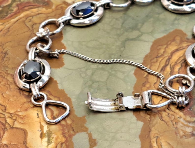 Op de afbeelding: Een zilveren armband met zwarte ovale stenen en een zilveren sluiting. De armband ligt op een bruine en witte achtergrond.