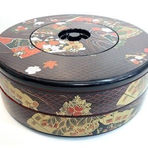 May include: A round, tiered Japanese lacquerware box with a black lid and base. The box is decorated with traditional Japanese motifs in red, gold, and white on a dark brown background. The lid has a central circular indentation.