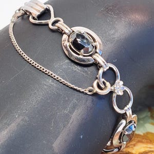 Op de afbeelding: Een zilveren armband met een ketting en zwarte ovale stenen. De armband heeft een decoratieve sluiting met een bloemmotief.