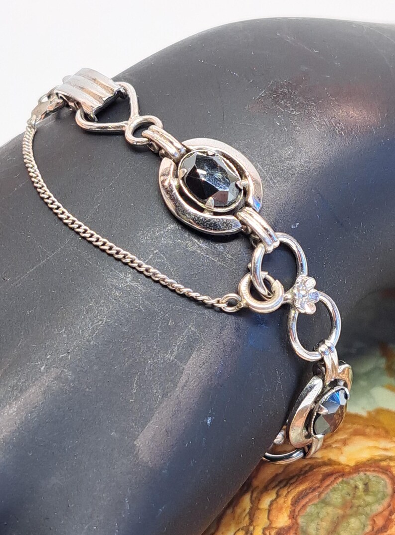 Op de afbeelding: Een zilveren armband met een ketting en zwarte ovale stenen. De armband heeft een decoratieve sluiting met een bloemmotief.