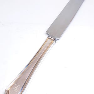 May include: A silver butter knife with a rounded blade and a decorative handle. The handle has an engraved design and a monogram. The knife is made of stainless steel and is designed for spreading butter or other soft foods.