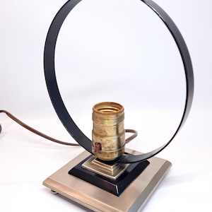 May include: A vintage table lamp with a black, circular shade and a square base. The base has a silver finish with black accents. The lamp features a brass bulb socket and a brown power cord. The lamp's design is mid-century modern.