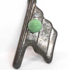 May include: A vintage metal tool with a weathered, dark gray finish and a prominent, smooth, light green spherical knob attached to its surface. The object features a long, straight edge, a curved side, and two circular holes punched through the metal.