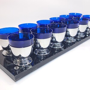 May include: A set of ten cobalt blue glass cups with silver-toned metal bases, arranged in two rows on a rectangular black marble-patterned tray. Each cup features a deep blue bowl set into a polished silver pedestal base with a circular foot.