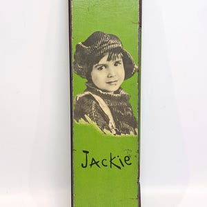 May include: A green rectangular box with a black border. The box has a black and white photo of a young girl wearing a hat and a sweater. The text "Jackie" is written in black cursive on the box.