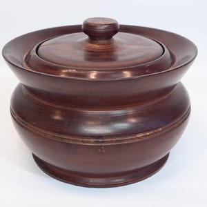 May include: A dark brown wooden lidded jar with a smooth, rounded body and a wide, flared rim. The lid features a small, centered knob handle. The wood grain is visible throughout the surface, showing a polished finish with subtle horizontal lines.