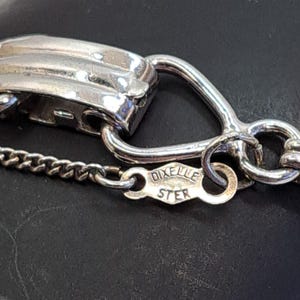Op de afbeelding: Close-up van een zilveren kettinghalsband sluiting met een 'DIXIE STER' stempel. De ketting is gemaakt van kleine, met elkaar verbonden zilveren stukjes.