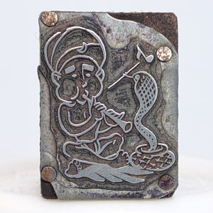 May include: A vintage metal printing block featuring a detailed illustration of a person playing a musical instrument while sitting on a snake. The block has a rustic, aged appearance with visible wear and tear.