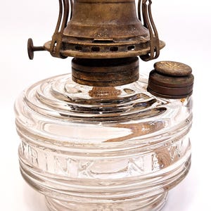 May include: A clear glass oil lamp base with horizontal ribbed detailing and the embossed text DRINK NEW. The lamp features a tarnished brass burner assembly with a side adjustment knob and a separate circular brass filler cap on the top surface.