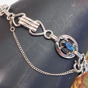 Op de afbeelding: Een zilveren armband met een blauwe strasssteen en een kettingdetail.
