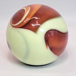 May include: A glass marble with a swirl of orange, brown, and white colors. The marble has a light green base color with a glossy finish. The design features a spiral pattern and a rounded shape.
