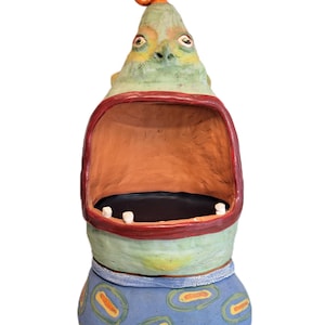 May include: A whimsical ceramic sculpture of a creature with a green face, orange swirl on top, and a large open mouth. The sculpture has a blue base with an oval pattern and a red rim around the mouth.
