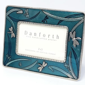 May include: A rectangular pewter picture frame with a teal enamel background. The frame is decorated with dragonfly accents and long, curved leaf-like designs. The frame has a 2x3 inch opening and the text "Danforth" is visible.