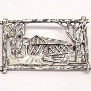 May include: Silver-toned brooch featuring a covered bridge scene within a rectangular frame. The design includes trees on either side and a detailed depiction of the bridge and surrounding landscape. A classic accessory.
