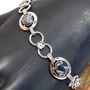 Op de afbeelding: Een zilveren armband met zwarte edelstenen en bloemmotieven. De armband heeft een kettingontwerp met ronde en ovale schakels.