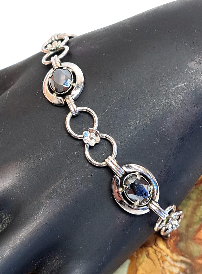 Op de afbeelding: Een zilveren armband met zwarte edelstenen en bloemmotieven. De armband heeft een kettingontwerp met ronde en ovale schakels.