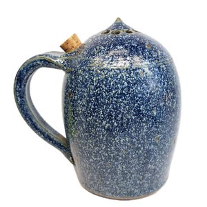 May include: A speckled blue ceramic pitcher with a handle and a cork stopper. The pitcher has a rounded body and a pointed top with several small holes. The handle is curved and attached to the side of the pitcher.