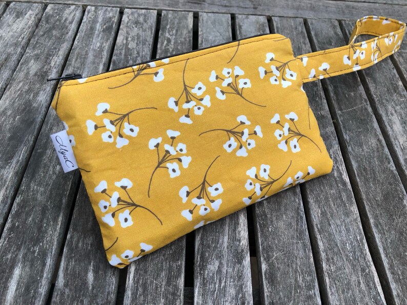 Puede incluir: Bolsa con cremallera de color amarillo mostaza con un estampado floral blanco y un asa peque&ntilde;a. La bolsa tiene una cremallera negra y una peque&ntilde;a etiqueta blanca con el texto "Alysa C".