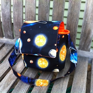 Pu&ograve; includere: Una borsa in tessuto strutturato con sfondo nero e una stampa spaziale colorata raffigurante soli gialli, pianeti blu e stelle bianche. La borsa &egrave; dotata di una patta, una tracolla regolabile e una fodera interna di colore arancione brillante.