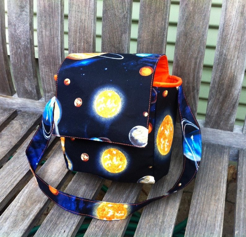 Pu&ograve; includere: Una borsa in tessuto strutturato con sfondo nero e una stampa spaziale colorata raffigurante soli gialli, pianeti blu e stelle bianche. La borsa &egrave; dotata di una patta, una tracolla regolabile e una fodera interna di colore arancione brillante.