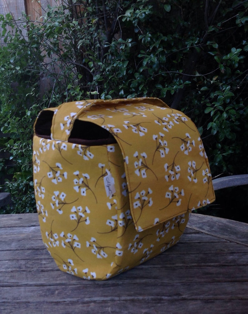 May include: A mustard yellow bag with a white floral pattern. The bag has a flap closure and a brown interior. The bag is sitting on a wooden surface. The bag is a tote bag.